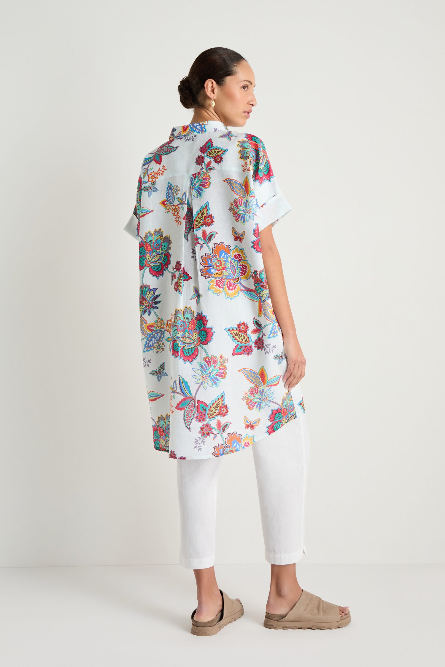 A woman stands sideways against a plain white background, wearing the Botanical Linen Long Shirt with white pants and beige sandals.