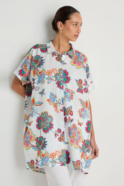 A woman models the Botanical Linen Long Shirt, featuring an oversized fit and vibrant botanical print, standing against a plain light background and gazing to the side.
