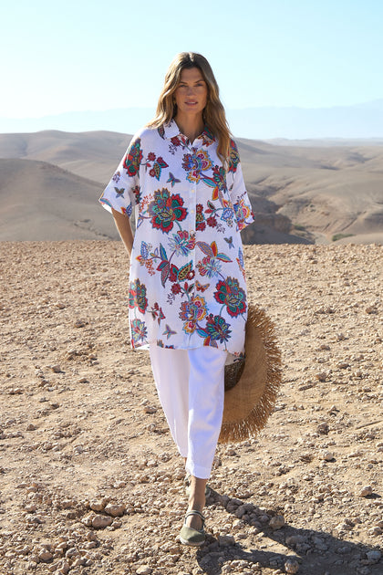 A woman stands in a rocky desert landscape wearing a Botanical Linen Long Shirt with an oversized fit, paired with white pants, flat shoes, and holding a large straw hat.