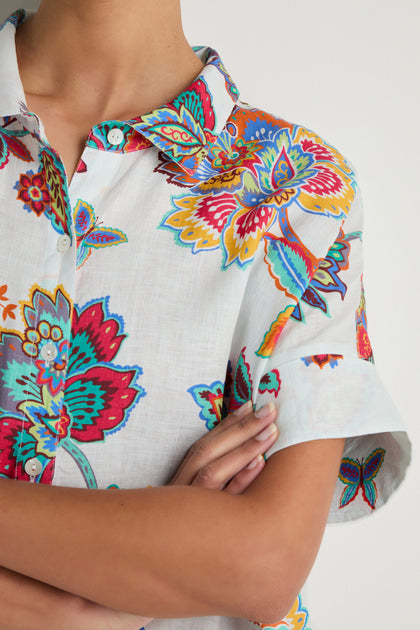 A person with arms crossed is wearing the Botanical Linen Long Shirt, featuring a colorful floral print on a white background. Only their upper torso and part of the face are visible.