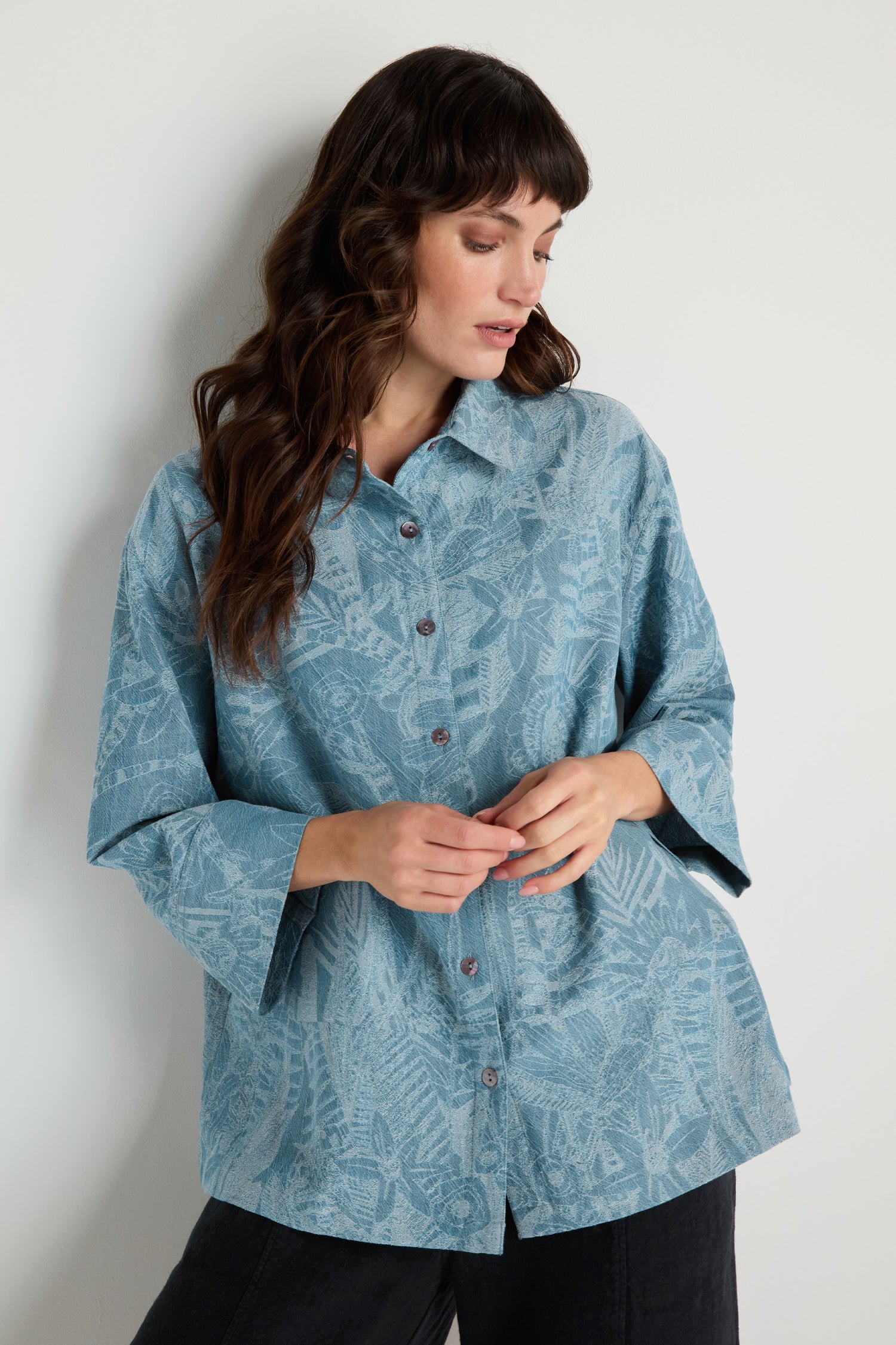 A woman with wavy brown hair wears the Floral Jacquard Cotton Shirt, a loose-fitting, boxy light blue top with a subtle leaf pattern, standing against a plain light-colored wall.
