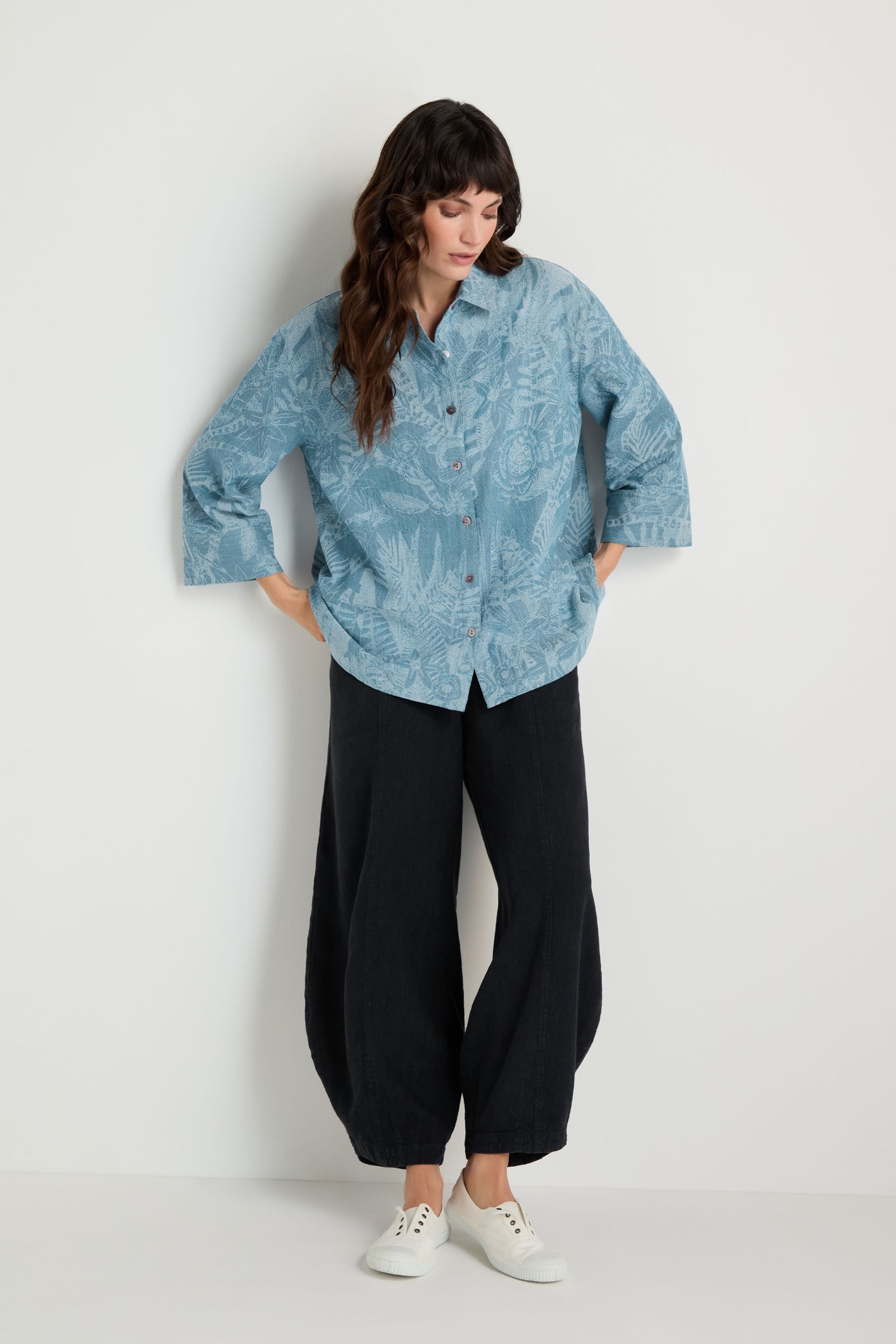 Woman standing against a white wall, wearing a Floral Jacquard Cotton Shirt with a boxy fit, loose black pants, and white sneakers; hands in her pockets and looking down.