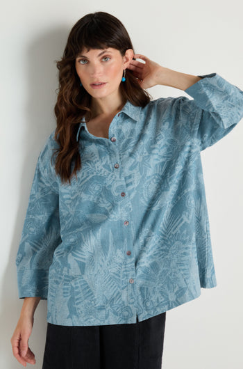 A woman stands against a white wall wearing the Floral Jacquard Cotton Shirt in classic denim blue with a boxy fit and black pants, raising one hand to her hair.