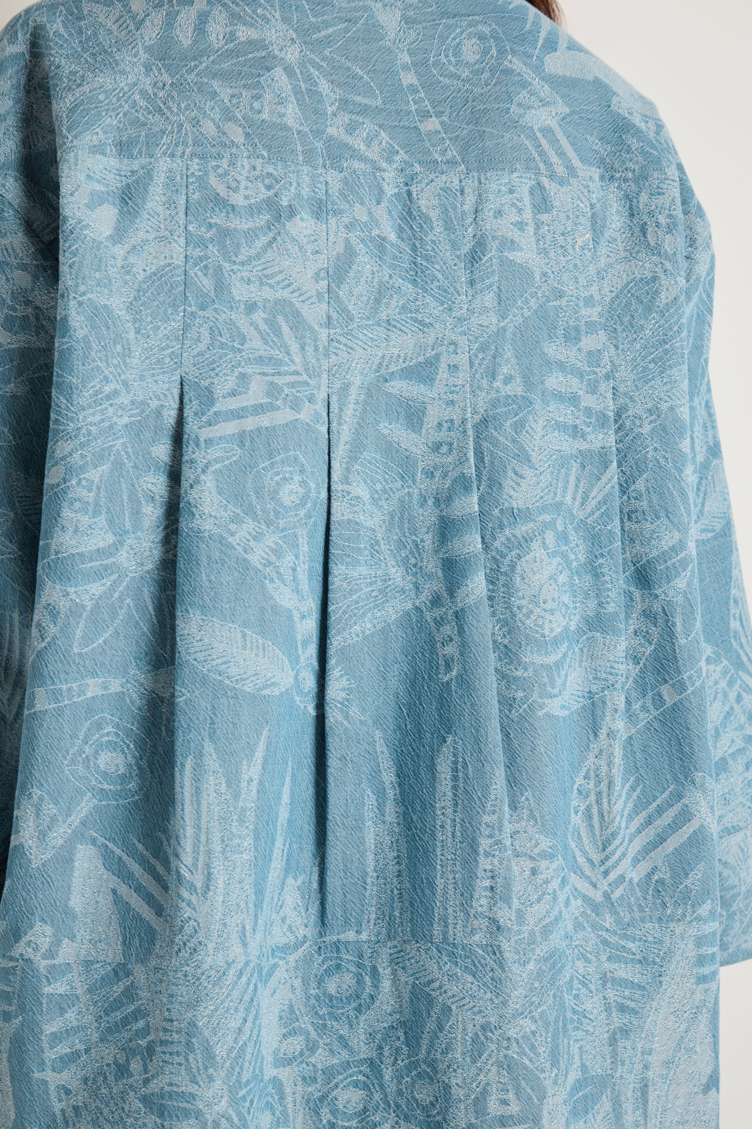 Close-up of the back of the Floral Jacquard Cotton Shirt, featuring a denim blue textured finish, abstract botanical pattern, visible pleats, and a boxy silhouette.