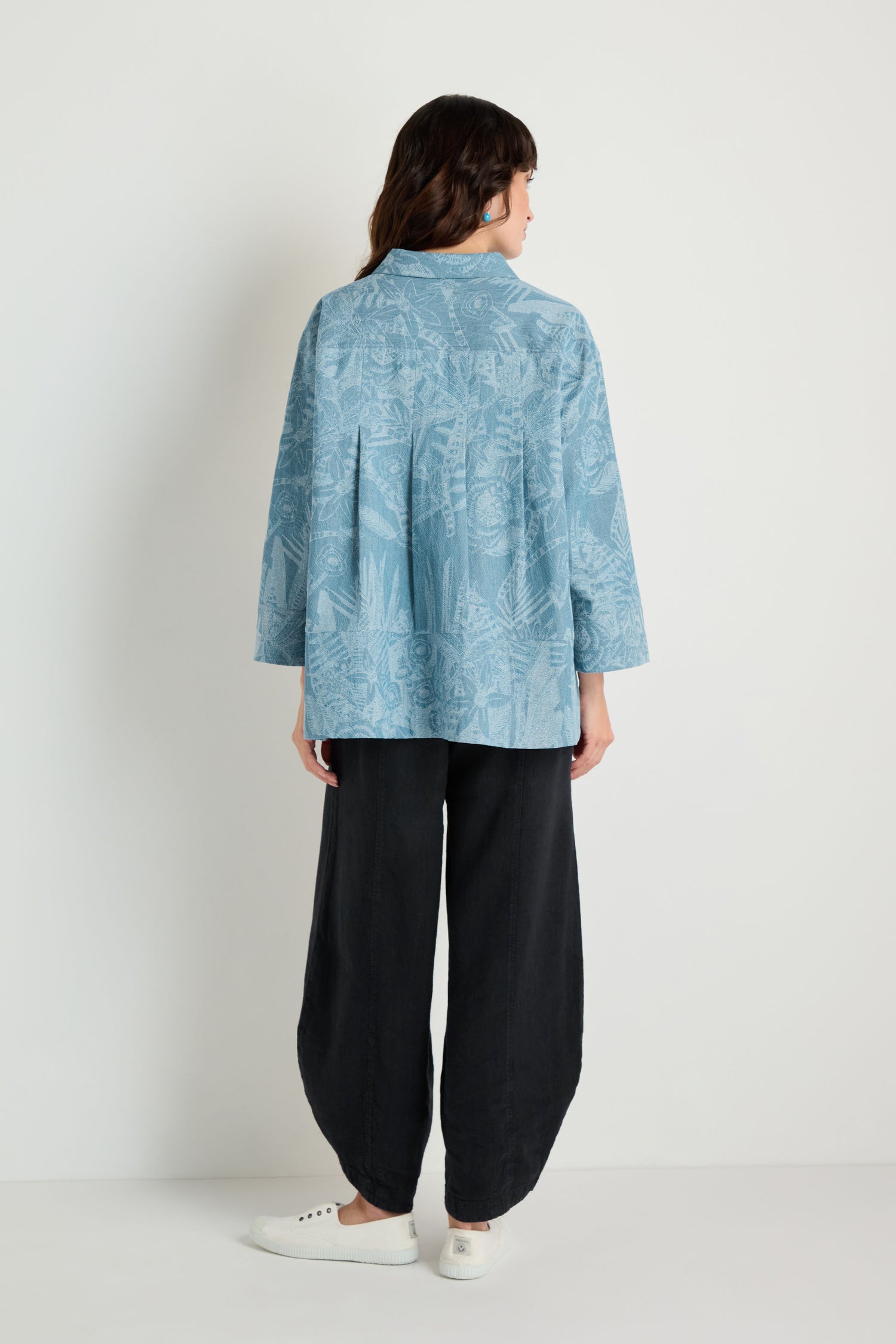 A woman, seen from behind against a plain white background, wears the Floral Jacquard Cotton Shirt in light blue, paired with loose black pants and white sneakers.
