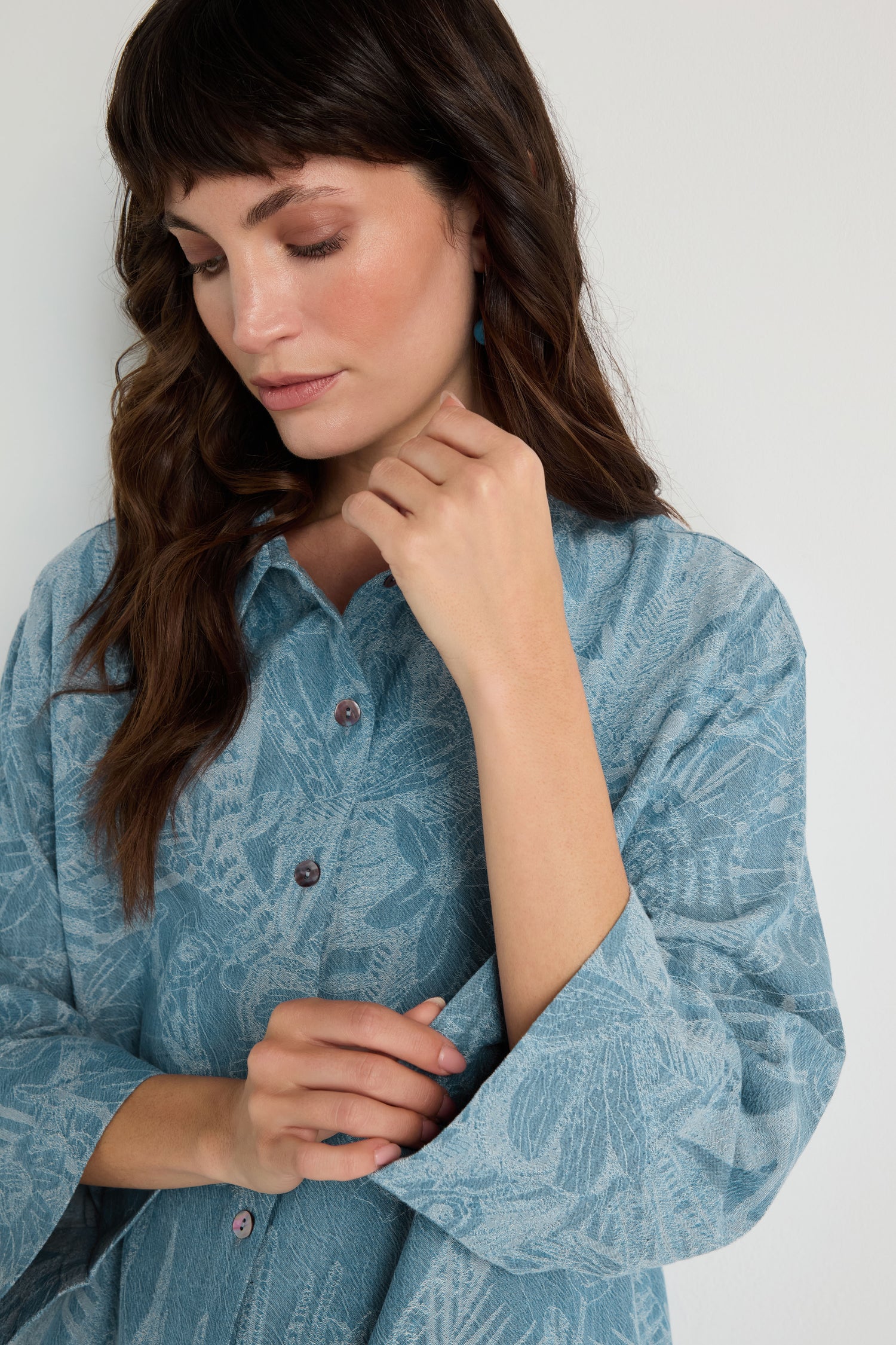 A woman with long brown hair wears the Floral Jacquard Cotton Shirt, standing against a white background and looking down with one hand touching her collar.