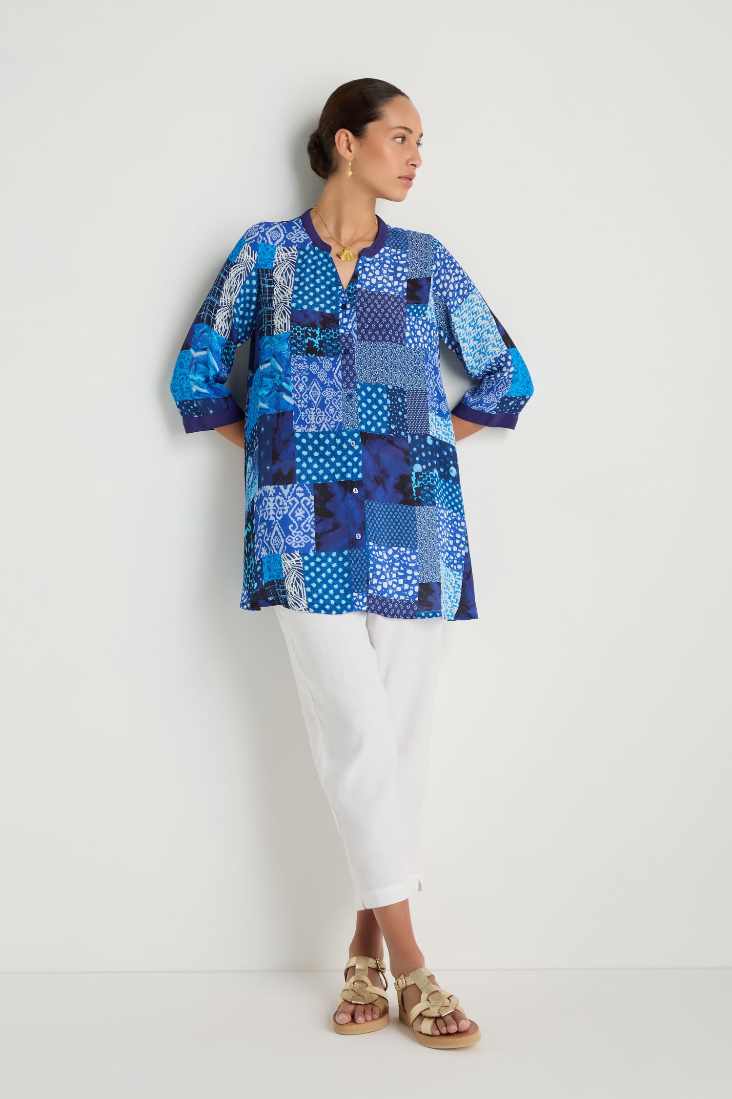 A woman stands against a white wall, wearing the Sahara Indigo Patchwork Longline Shirt in soft viscose crepe, paired with white pants and beige sandals, arms relaxed and head turned to the side.