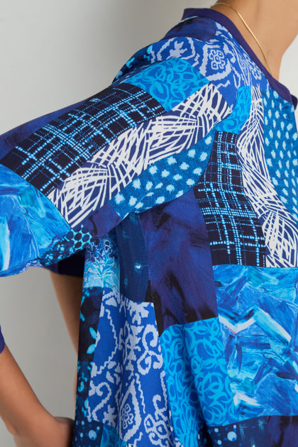 Model wearing the Sahara Indigo Patchwork Longline Shirt, a blue patterned viscose crepe piece with geometric and abstract designs, shown in profile with bent arm and hand on hip.