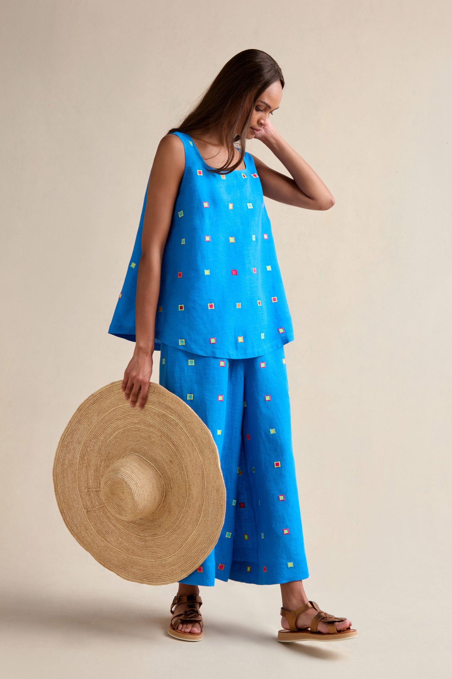 Woman in a relaxed fit Embroidered Squares Linen Vest, blue with small colorful square patterns, paired with matching wide-leg pants, holding a straw hat and wearing sandals.
