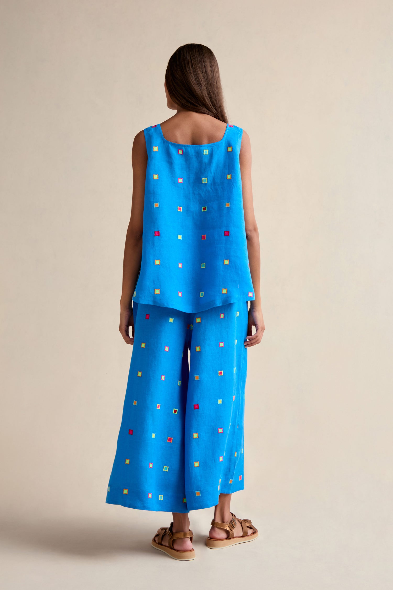 A woman faces away, wearing the Embroidered Squares Linen Vest in blue, paired with matching wide-leg pants featuring multicolored square patterns and brown sandals.