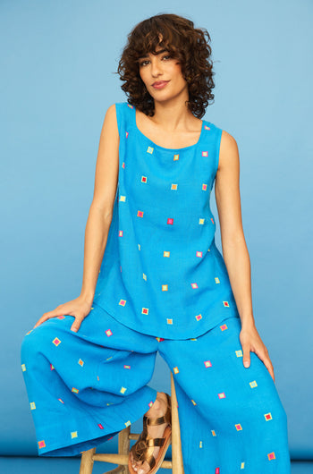 A woman with curly hair sits on a wooden stool, wearing the Embroidered Squares Linen Vest and matching wide-leg pants featuring colorful square patterns, set against a blue background.