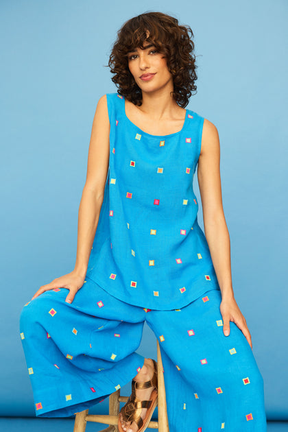 A woman with curly hair sits on a wooden stool, wearing the Embroidered Squares Linen Vest and matching wide-leg pants featuring colorful square patterns, set against a blue background.