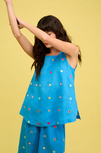 Woman wears the Embroidered Squares Linen Vest and matching pants with multicolored square patterns, standing against a yellow background with her arms raised.