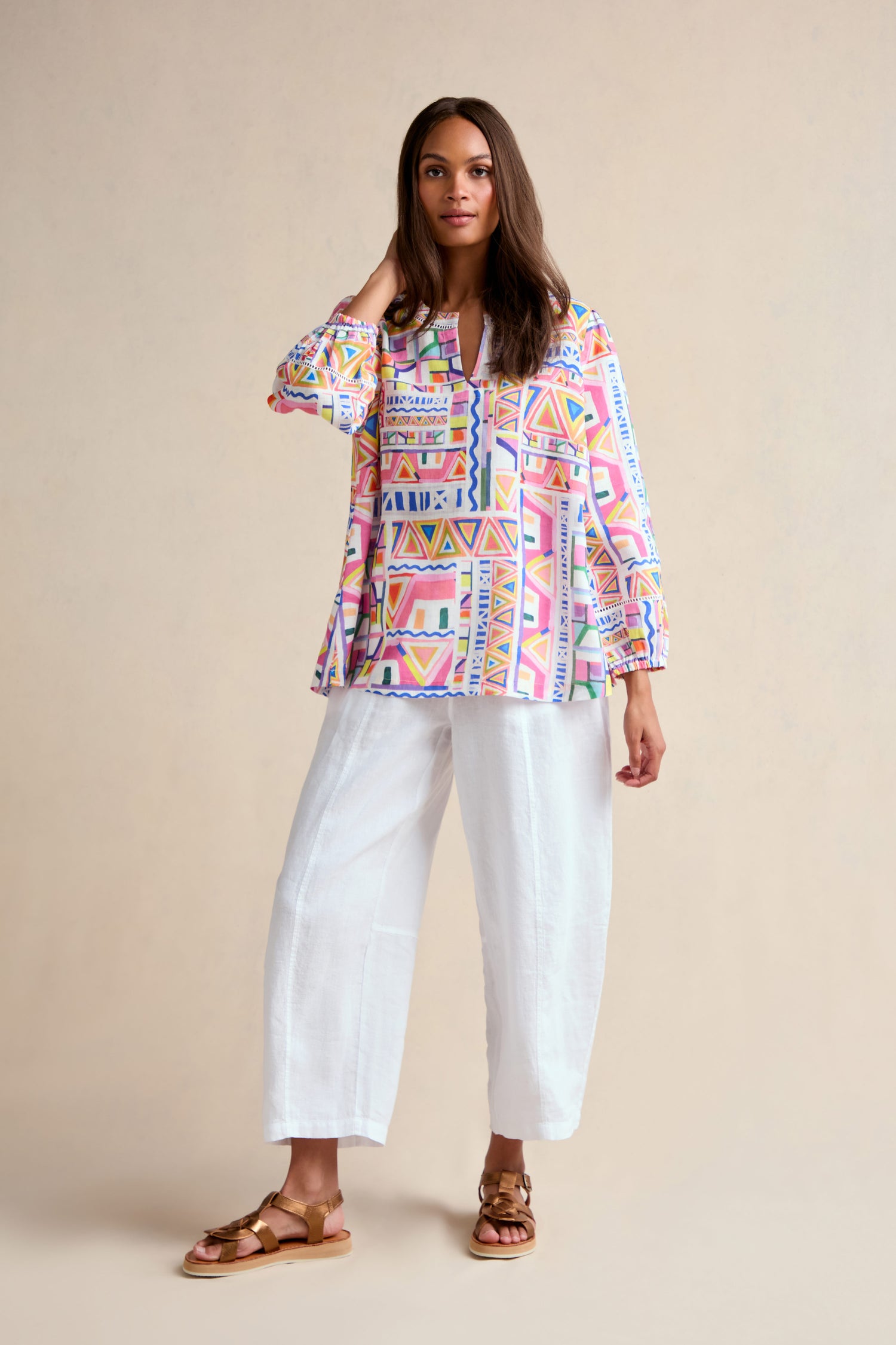 A woman stands against a plain background, wearing the Painted Geo Linen Top with balloon sleeves, paired with loose white pants and brown sandals.