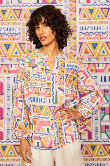 A woman with curly hair wears the Painted Geo Linen Top, featuring geometric patterns and balloon sleeves, paired with white pants, standing before a matching geometric-patterned background.