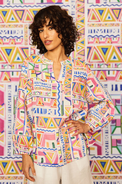 A woman with curly hair wears the Painted Geo Linen Top, featuring geometric patterns and balloon sleeves, paired with white pants, standing before a matching geometric-patterned background.
