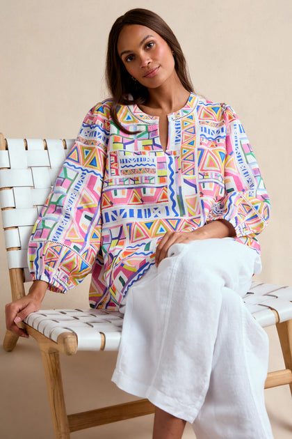 A woman sits on a wooden chair wearing the Painted Geo Linen Top, featuring colorful geometric patterns and balloon sleeves, paired with loose white pants as she gazes slightly to the side.