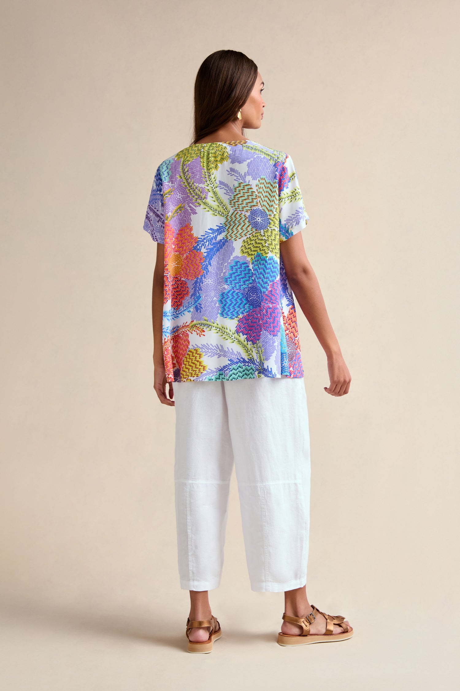A woman faces away, wearing the Pixelated Petals Print Top with a relaxed A-line fit, short sleeves, and a pixelated floral pattern. She pairs it with white cropped pants and brown sandals against a neutral background.