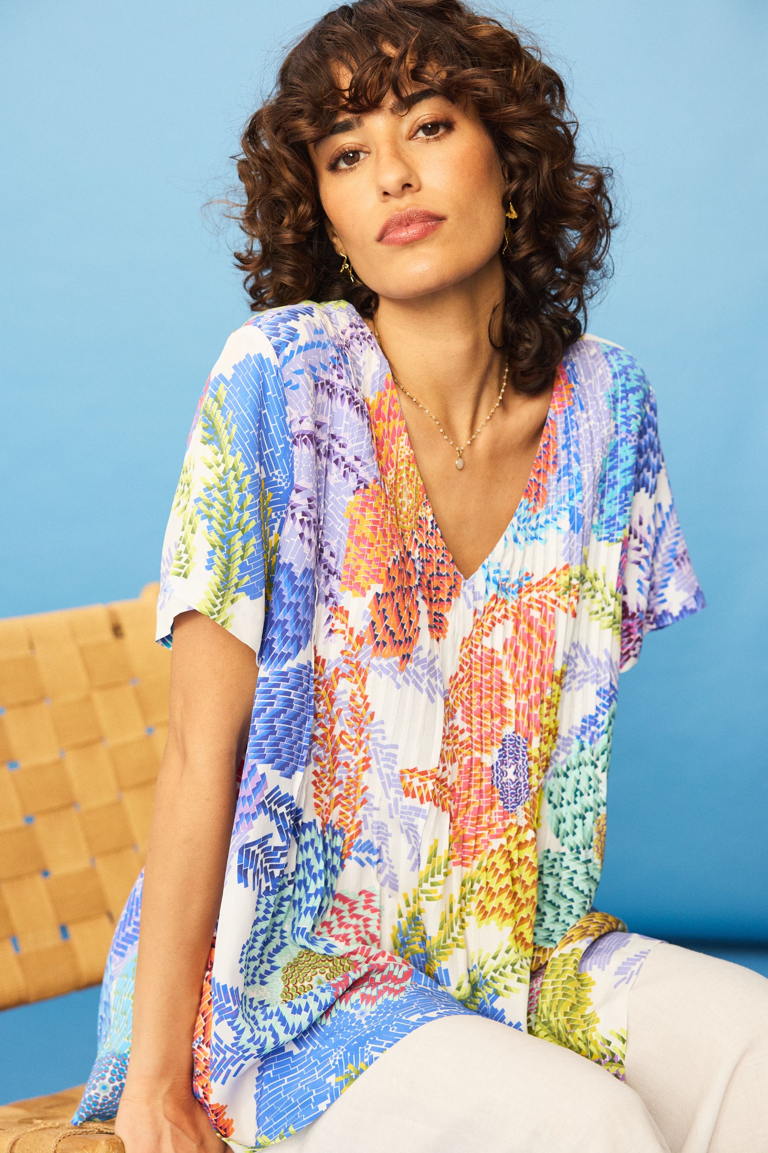 A woman with curly hair wears the Pixelated Petals Print Top—colorful, floral, short-sleeved, relaxed A-line fit—with white pants, sitting on a woven chair against a blue background.