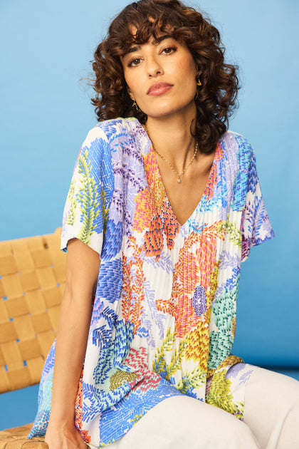 A woman with curly hair wears the Pixelated Petals Print Top—colorful, floral, short-sleeved, relaxed A-line fit—with white pants, sitting on a woven chair against a blue background.
