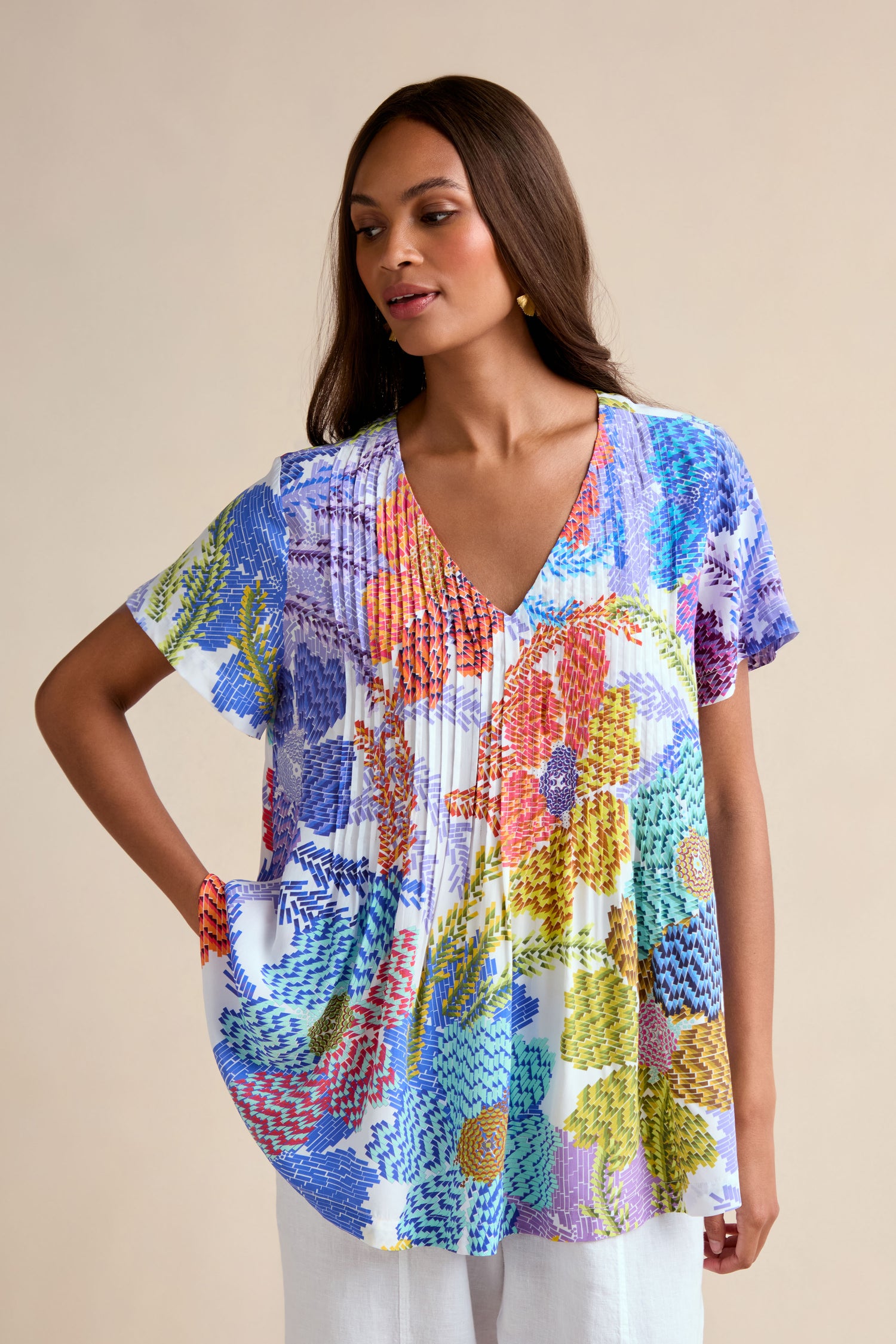 A woman models the Pixelated Petals Print Top—a relaxed A-line, short-sleeve blouse with pixelated floral print—paired with white pants, standing against a plain beige background.