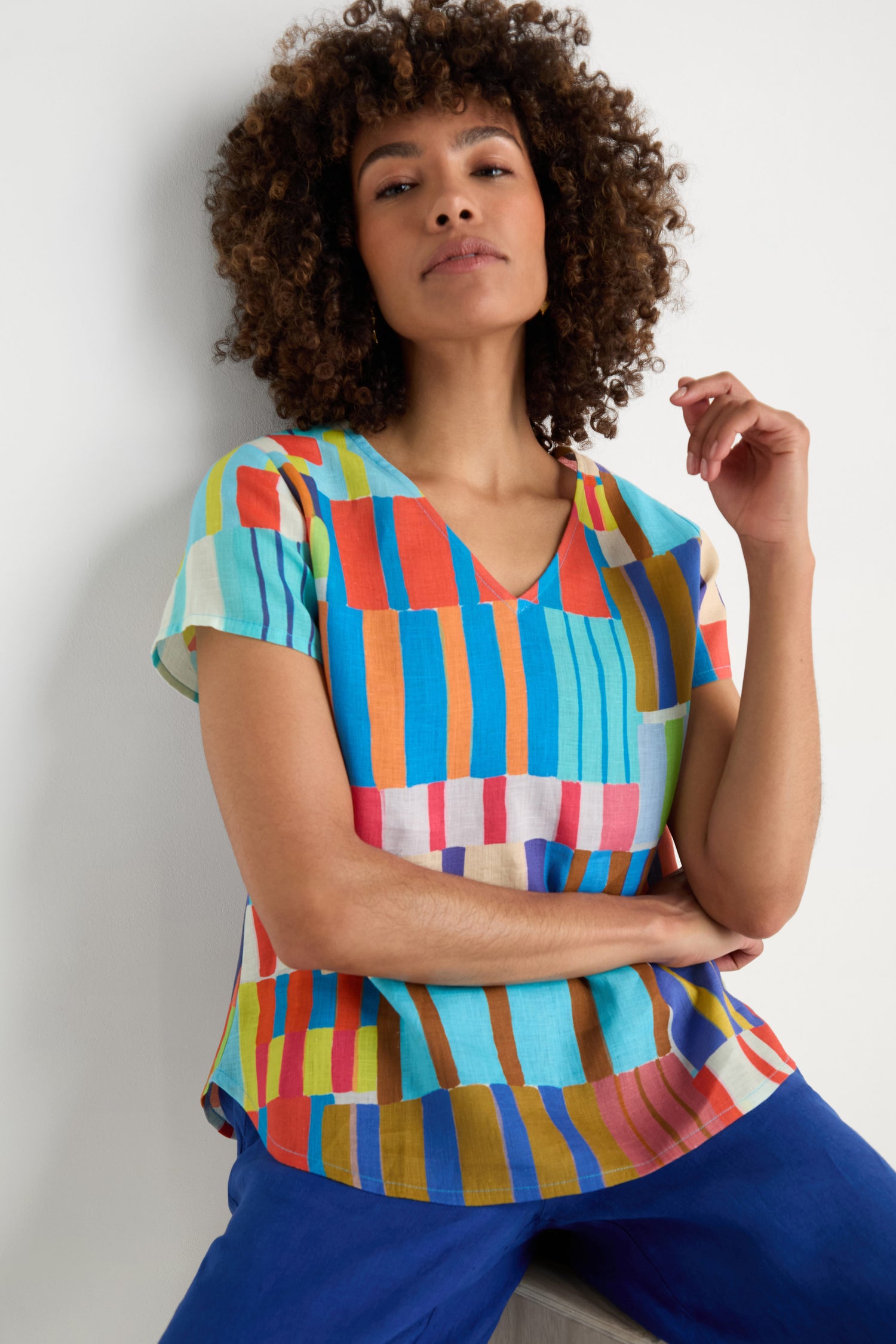 A person with curly hair wears the Painted Stripes Linen Top and blue pants, sitting casually against a white background.