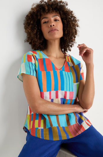 A person with curly hair wears the Painted Stripes Linen Top and blue pants, sitting casually against a white background.