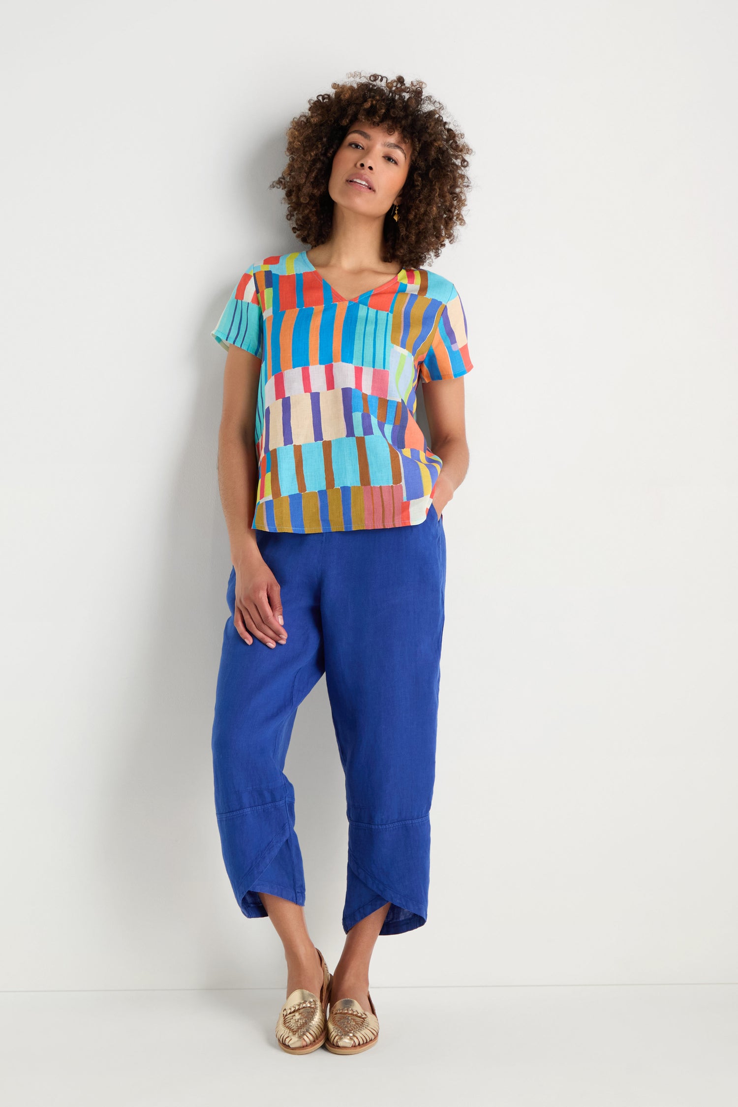 A woman stands against a white wall wearing the Painted Stripes Linen Top, blue cropped pants, and gold shoes with her hands in her pockets.