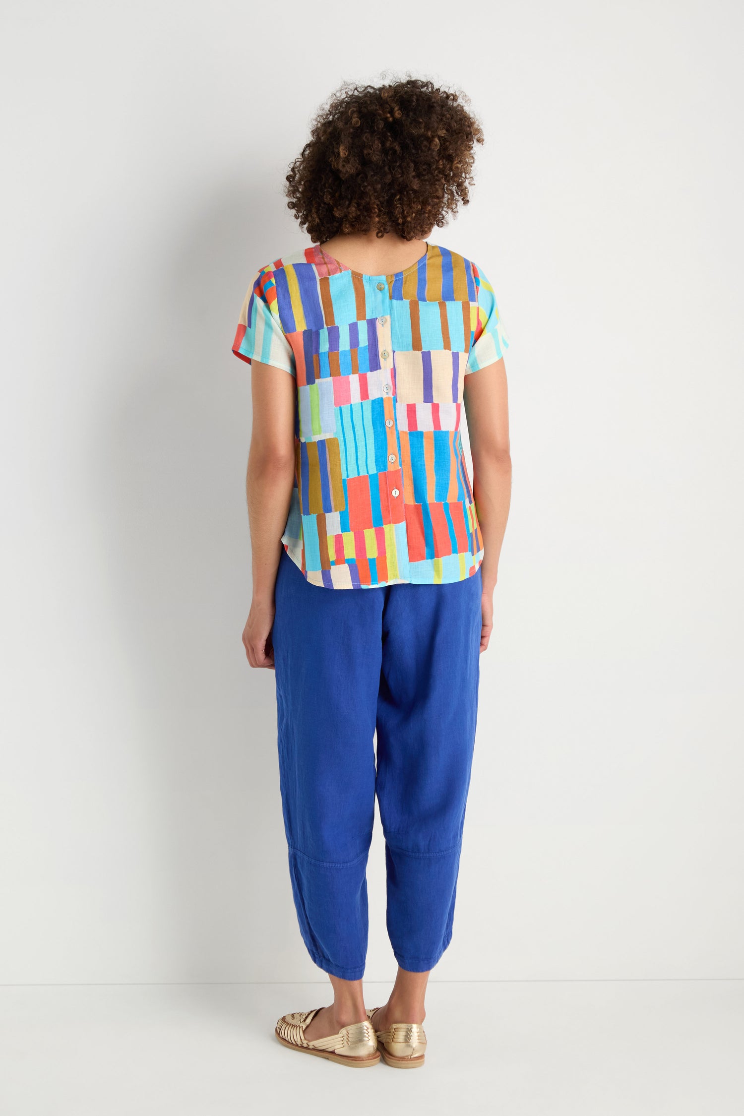 A person stands facing away, wearing the Painted Stripes Linen Top—colorful and relaxed fit—paired with blue pants and gold sandals, set against a plain white background.