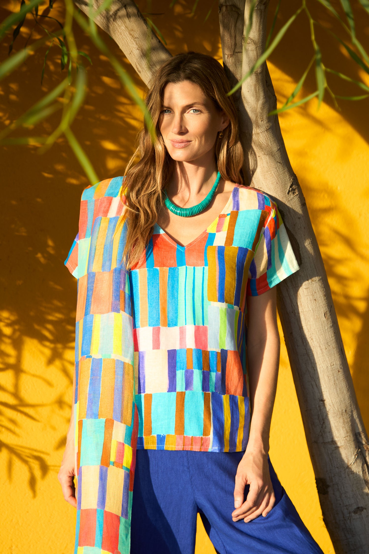 A woman stands by a tree against a yellow wall, wearing the vibrant Painted Stripes Linen Top with blue pants and a green necklace, sunlight casting striking shadows.