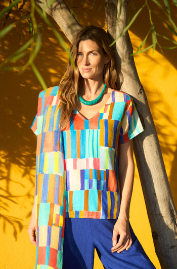 A woman stands by a tree against a yellow wall, wearing the vibrant Painted Stripes Linen Top with blue pants and a green necklace, sunlight casting striking shadows.