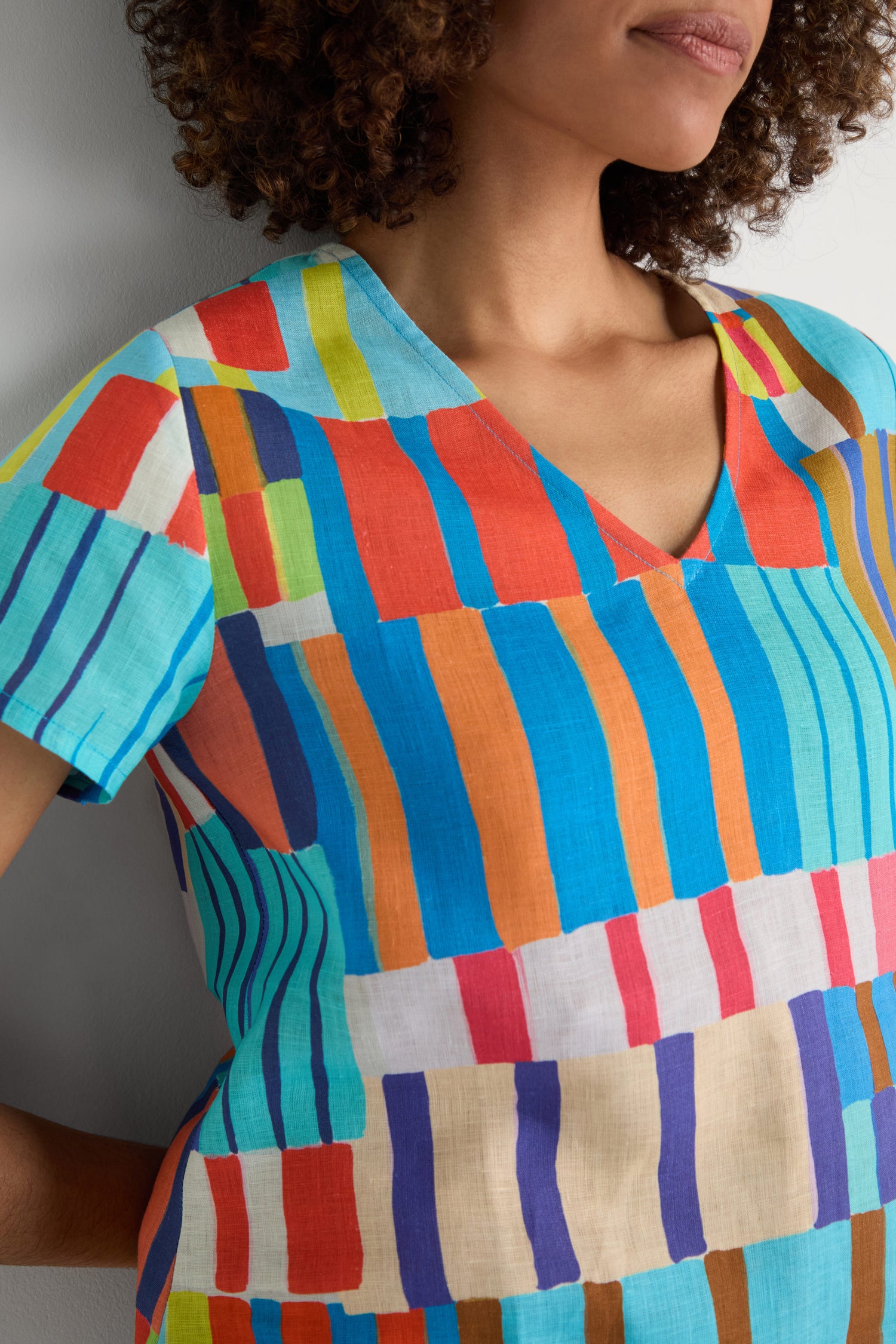 A person wears the Painted Stripes Linen Top, featuring a relaxed fit and bold, multicolored geometric patterns, standing against a light background with only their upper body and part of their face visible.