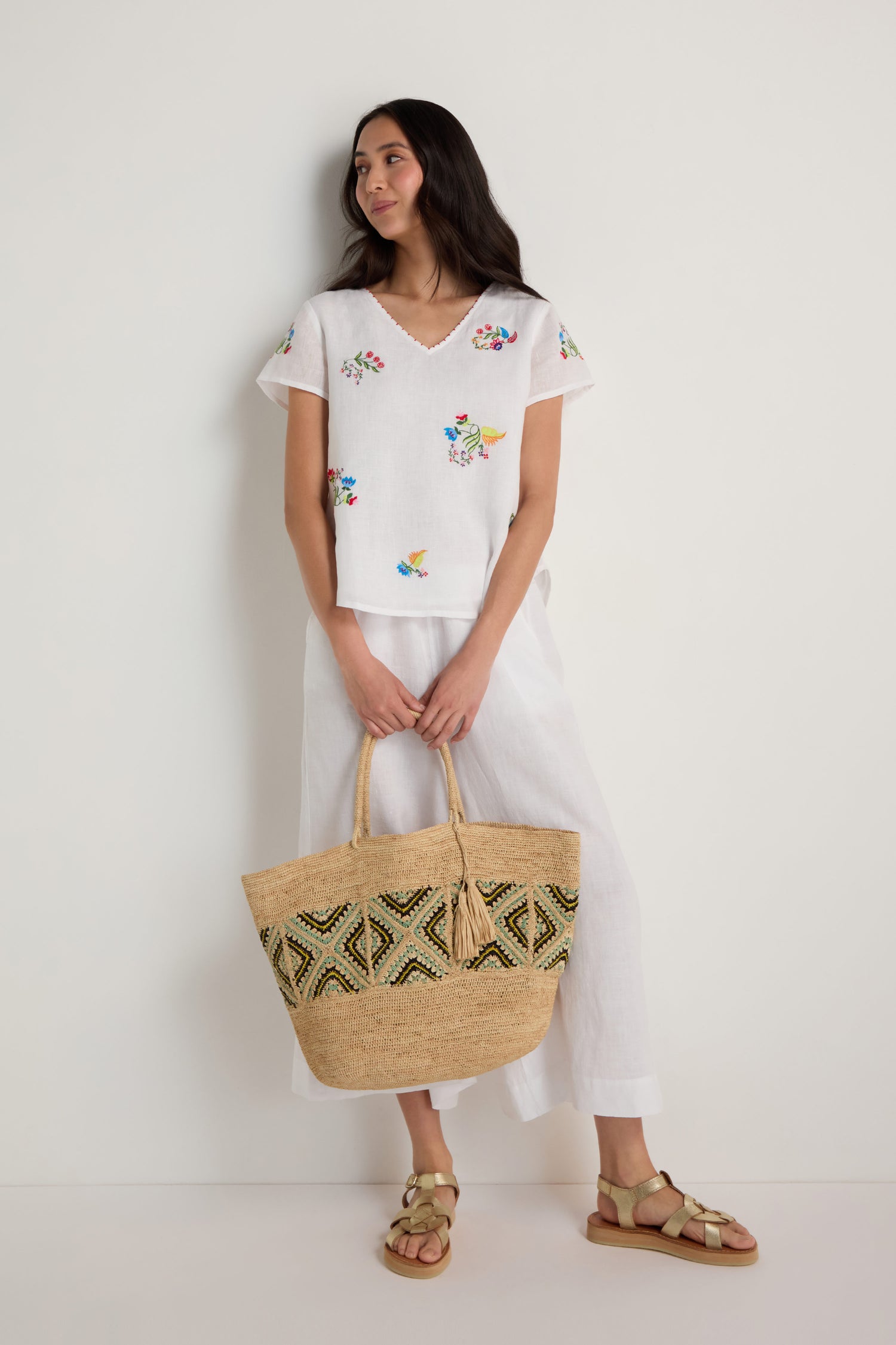 A woman stands against a white wall wearing a Vintage Floral Embroidered Top, white pants, tan sandals, and holding a large woven straw bag with a geometric pattern.