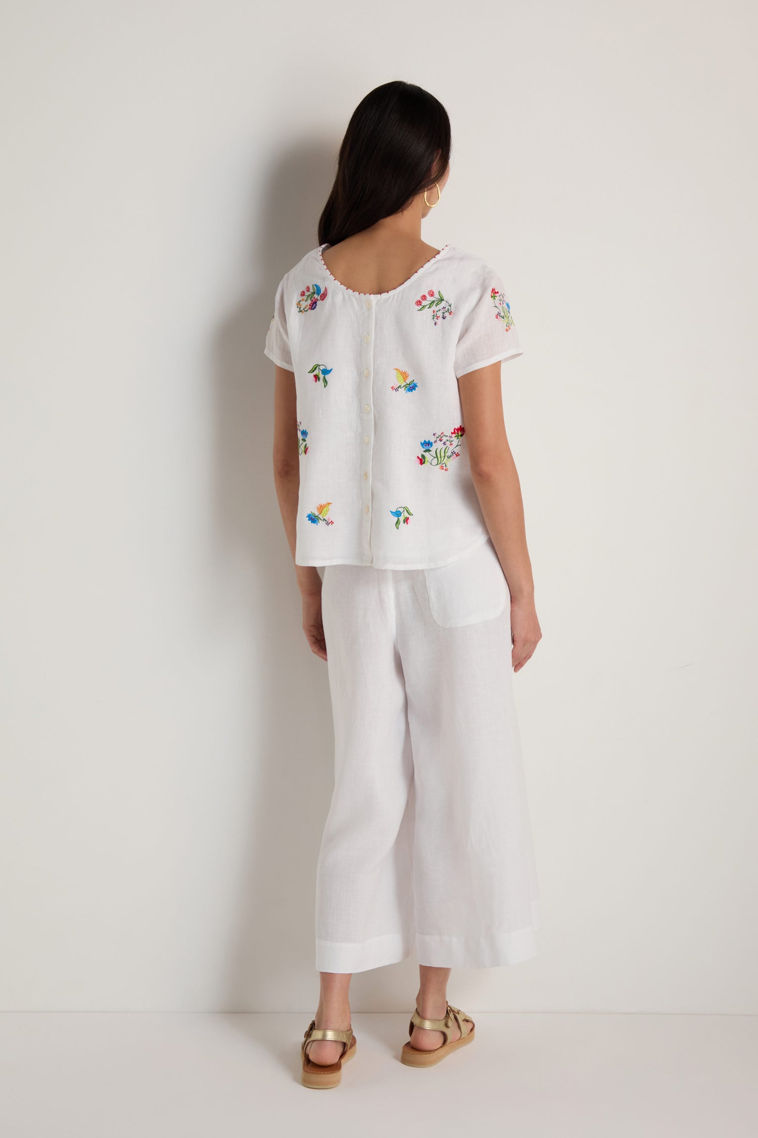 A woman stands against a white wall, facing away, wearing the Vintage Floral Embroidered Top paired with matching white wide-leg pants, sandals, and hoop earrings.