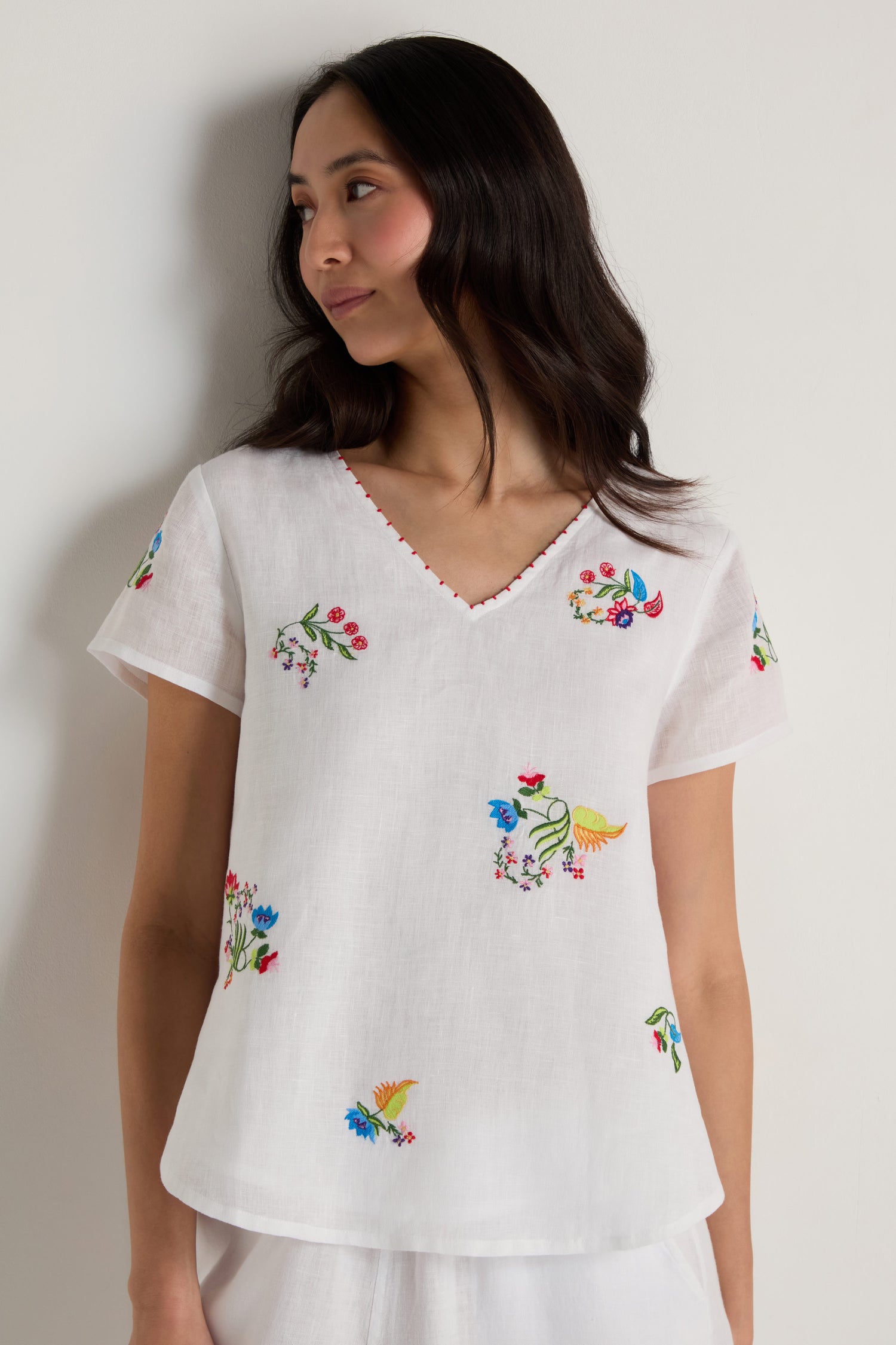 A woman wearing the Vintage Floral Embroidered Top, featuring white linen fabric, short sleeves, a V-neck, and colorful embroidered flowers, stands against a plain light background.
