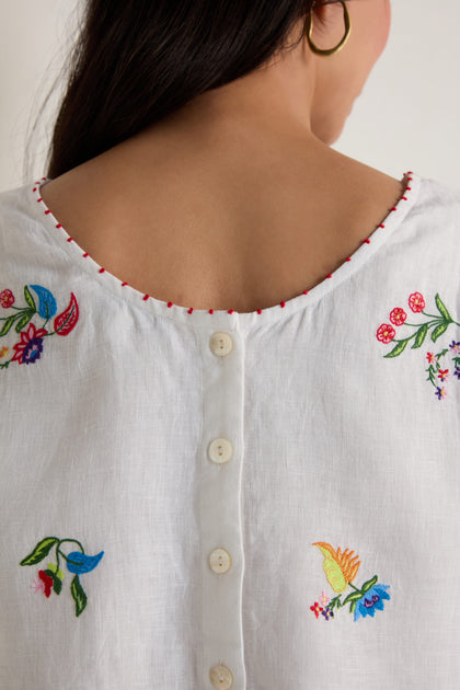 Close-up of a woman’s back in the Vintage Floral Embroidered Top—white linen featuring colorful floral embroidery and a row of white buttons down the back.