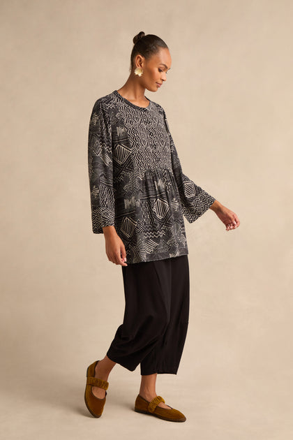 A woman stands sideways against a plain beige background, wearing the Mono Patchwork Ikat Tunic—crafted from soft viscose crepe in a patterned black and white design—paired with loose black pants and brown shoes.