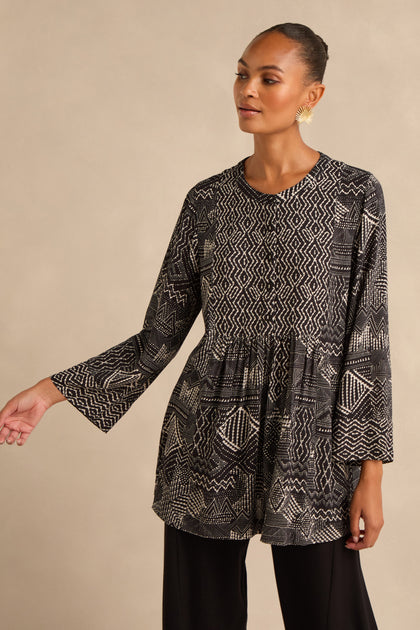 A woman wearing the Mono Patchwork Ikat Tunic, a long-sleeve black and white top featuring geometric patterns crafted from soft viscose crepe, paired with black pants, stands against a neutral background.
