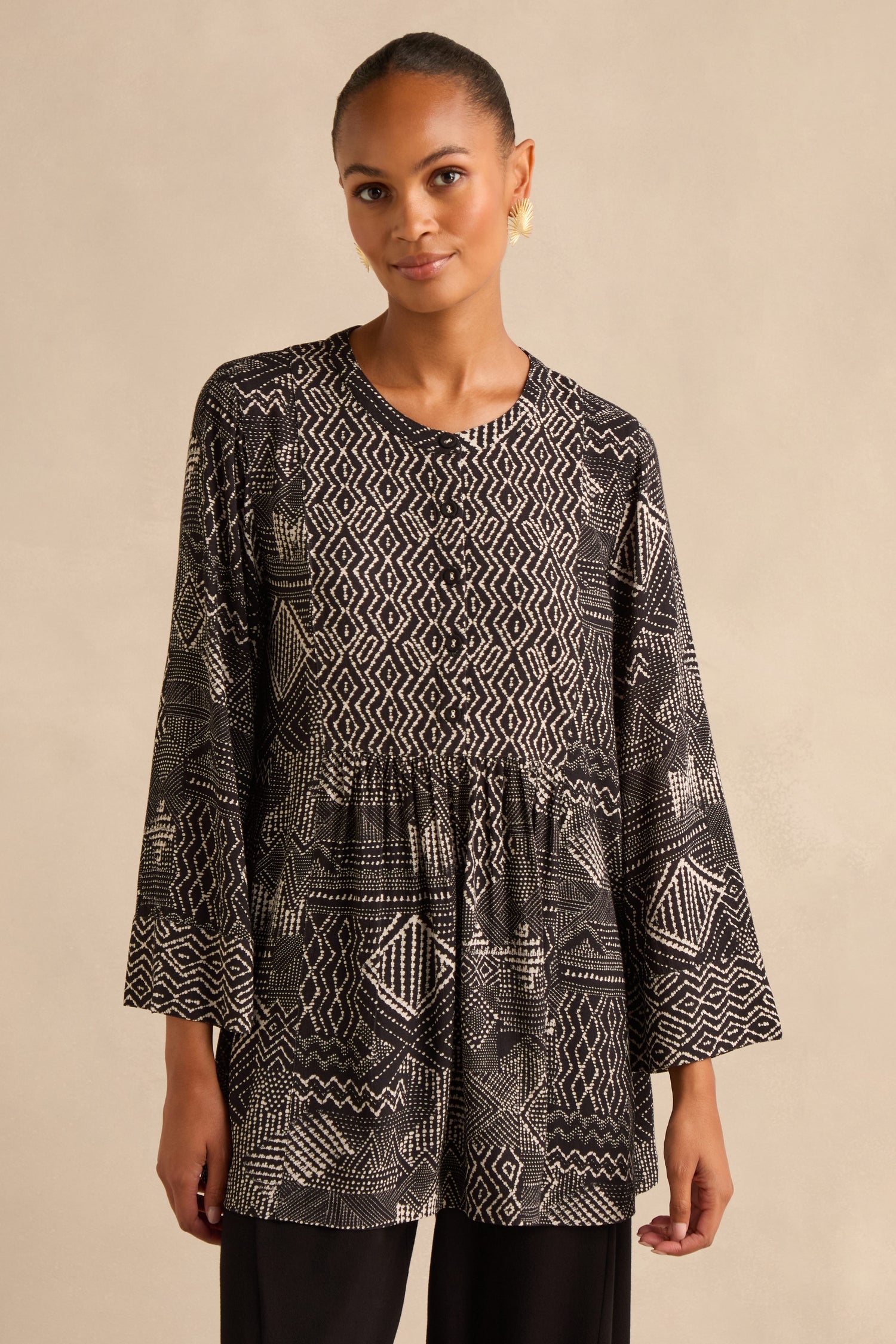 A woman wearing the Mono Patchwork Ikat Tunic—crafted from soft viscose crepe, featuring a loose, long-sleeved silhouette in black with white geometric patterns—stands against a neutral background.