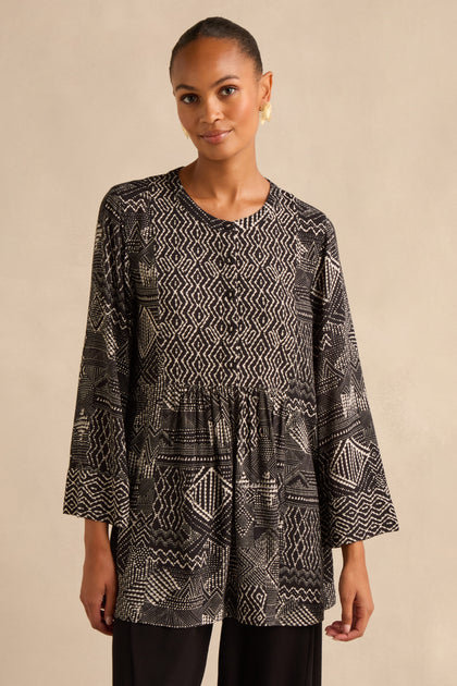 A woman wearing the Mono Patchwork Ikat Tunic—crafted from soft viscose crepe, featuring a loose, long-sleeved silhouette in black with white geometric patterns—stands against a neutral background.