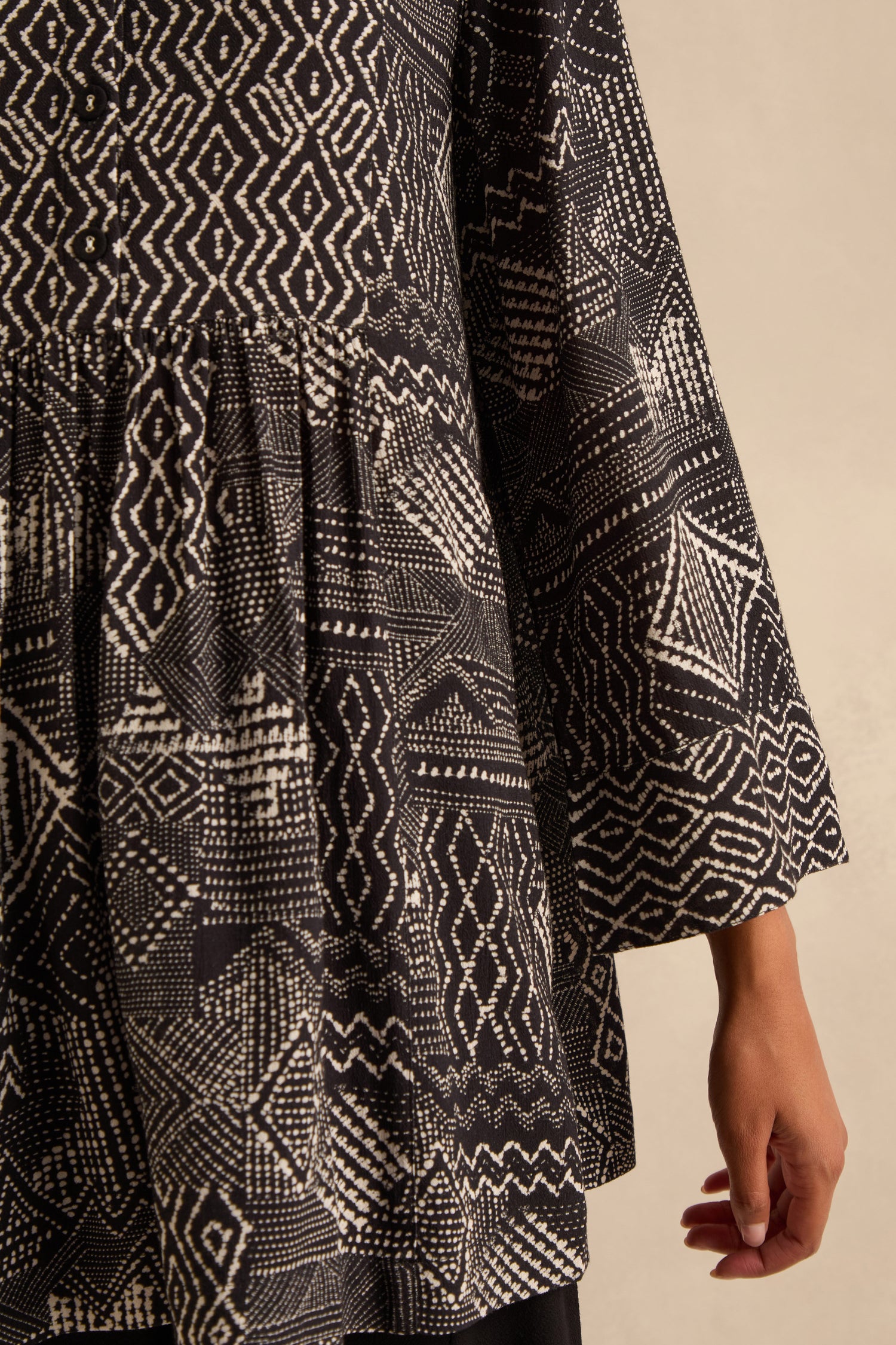 A person wearing the Mono Patchwork Ikat Tunic, a black and white patterned viscose crepe peasant style featuring geometric and dotted designs, is shown from the shoulders to mid-thigh.