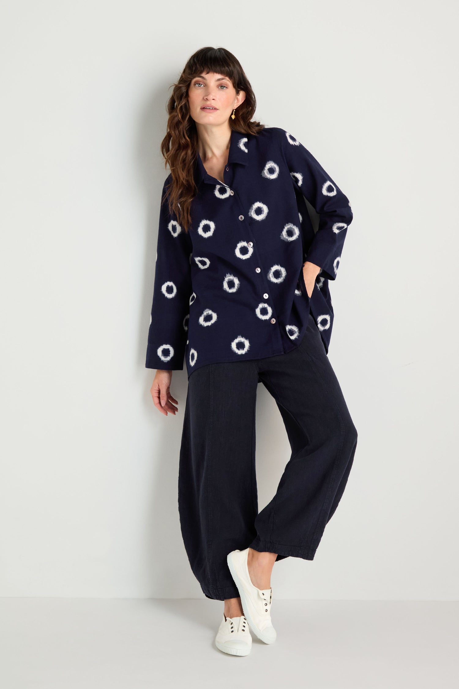 A person stands against a plain wall, wearing the Ikat Circles Cotton Shirt, wide-leg navy pants, and white sneakers.