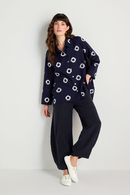 A person stands against a plain wall, wearing the Ikat Circles Cotton Shirt, wide-leg navy pants, and white sneakers.