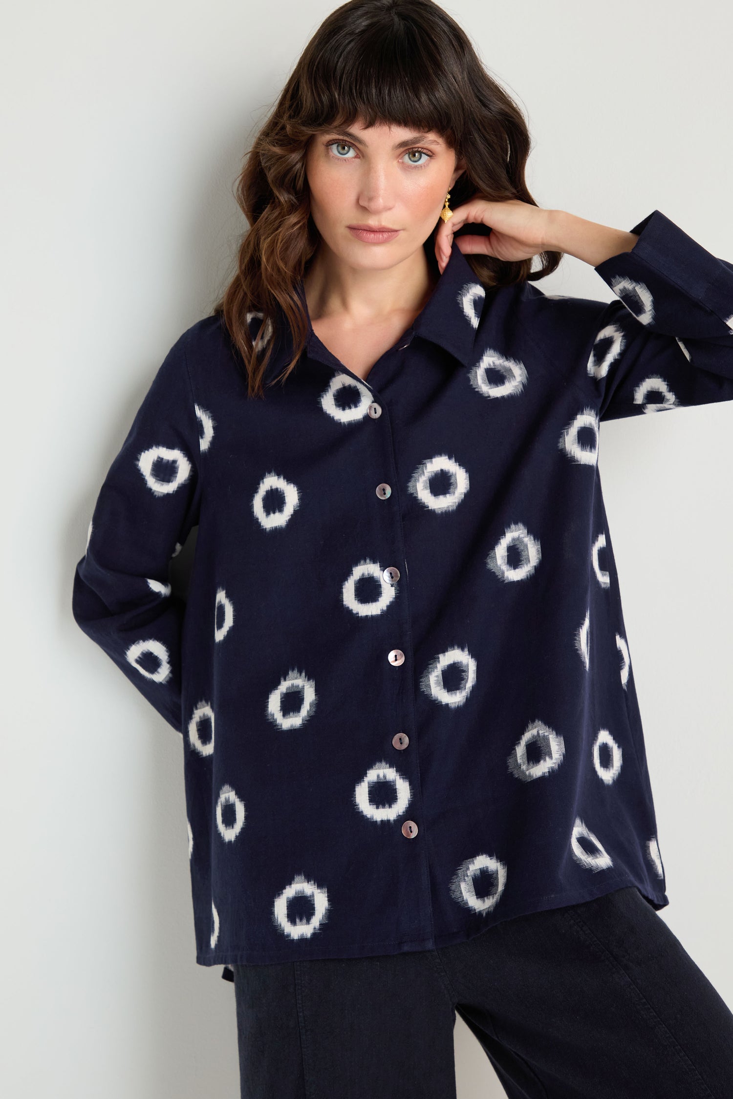 A woman stands against a light wall wearing the Ikat Circles Cotton Shirt in navy blue with white circular patterns, paired with dark pants.