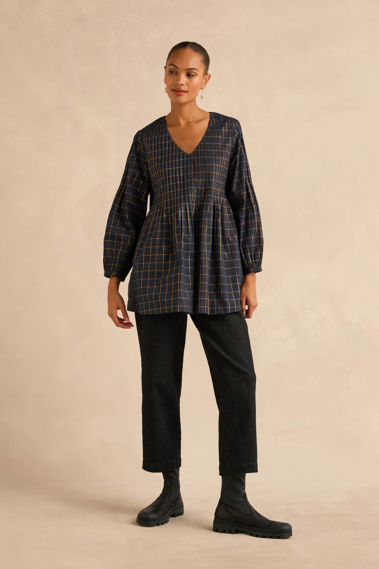 A person wears a dark navy Lurex Check Pintuck Tunic with a V-neck, pintuck detailing, and voluminous sleeves, paired with black trousers and black lug-sole ankle boots.