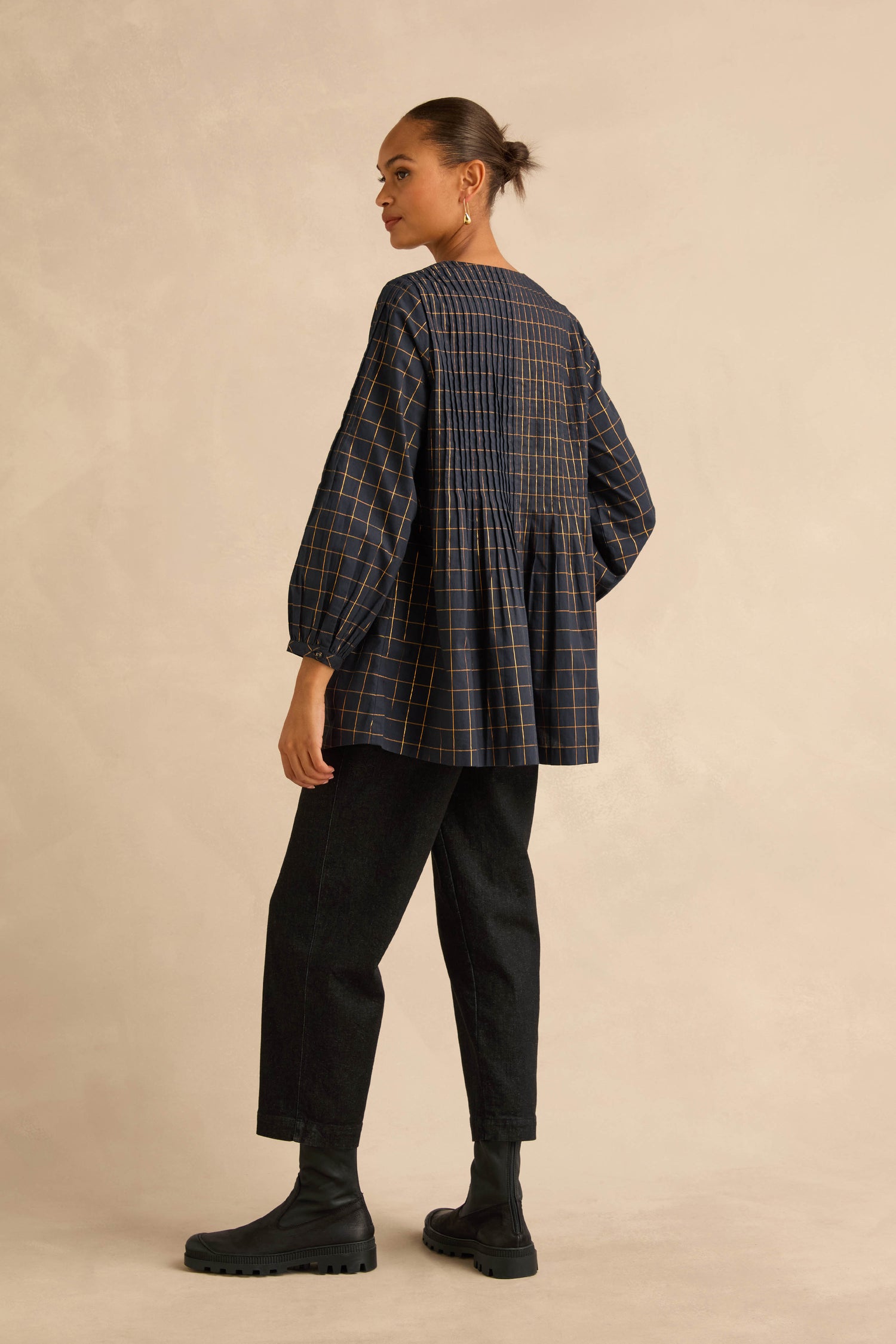 A woman wears a Lurex Check Pintuck Tunic in dark navy blue with a gold grid pattern and vertical pintuck pleats, paired with black trousers and ankle boots.