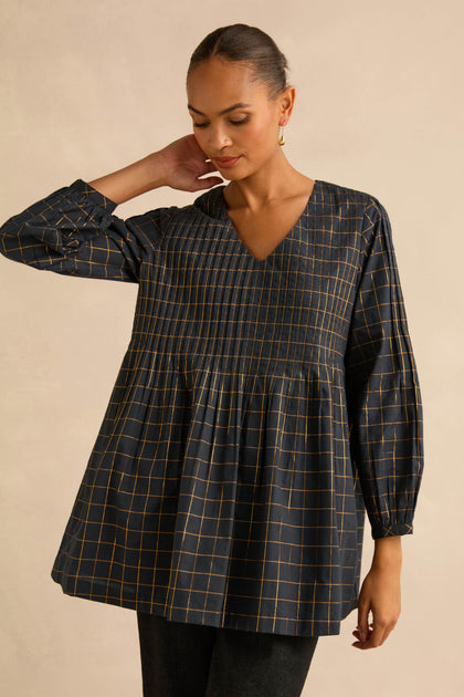 A woman wears a Lurex Check Pintuck Tunic in navy blue with a gold grid pattern, V-neck, vertical pintuck detailing, and a fit-and-flare silhouette.
