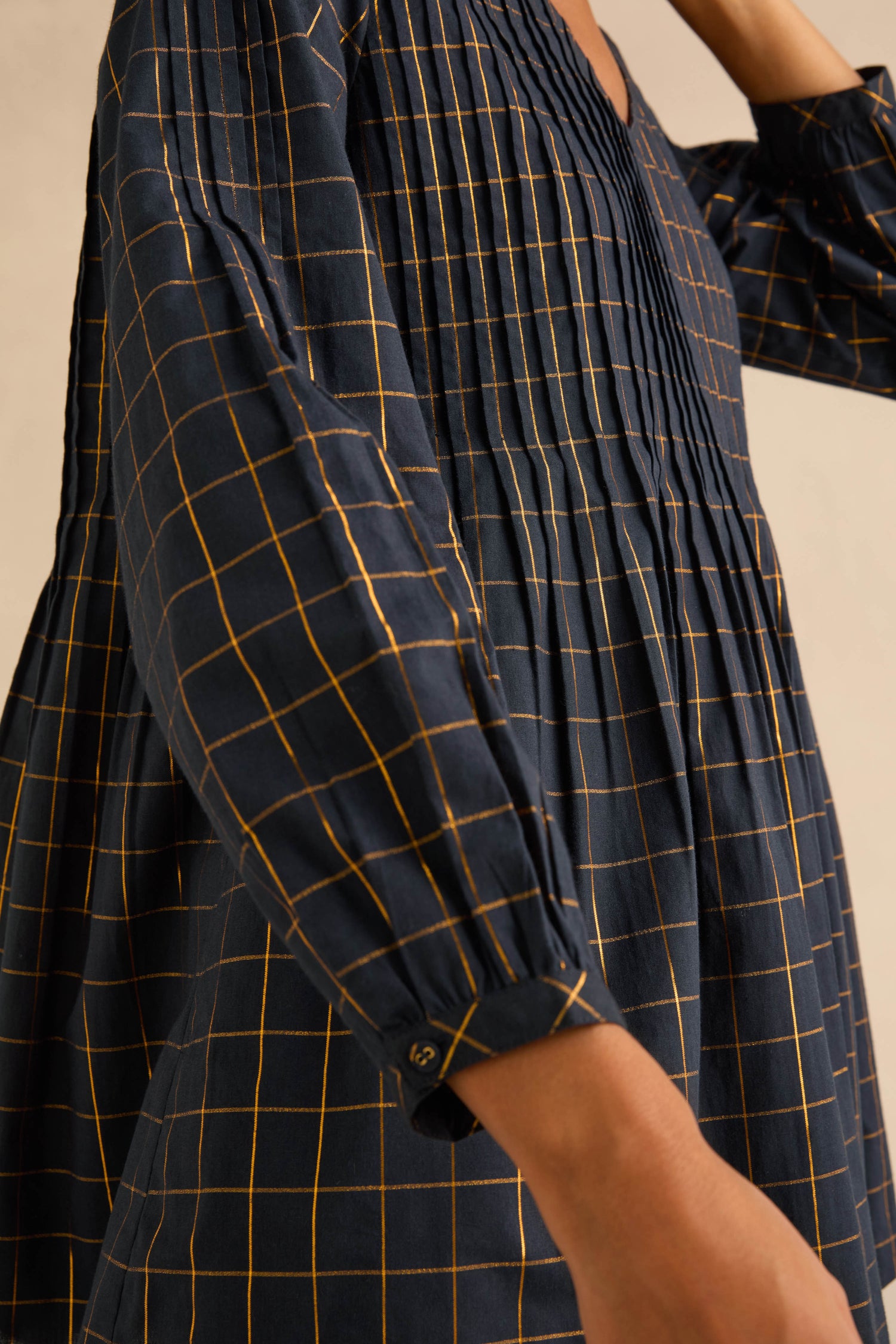 A person wearing a Lurex Check Pintuck Tunic in dark navy blue with a fit-and-flare silhouette, gold grid pattern, and vertical pintuck pleats at the bodice and sleeves.