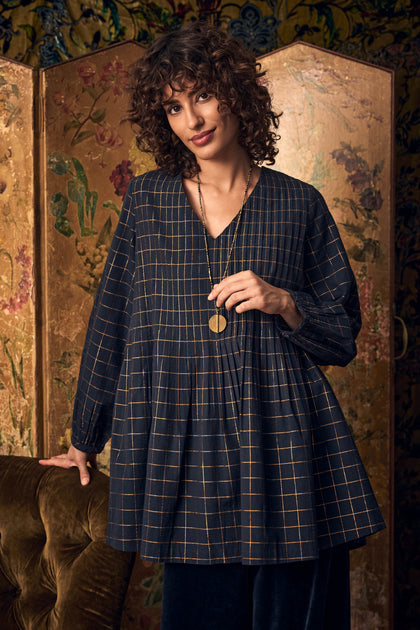 A person wearing a dark blue Lurex Check Pintuck Tunic with a metallic gold grid pattern and long pendant necklace.