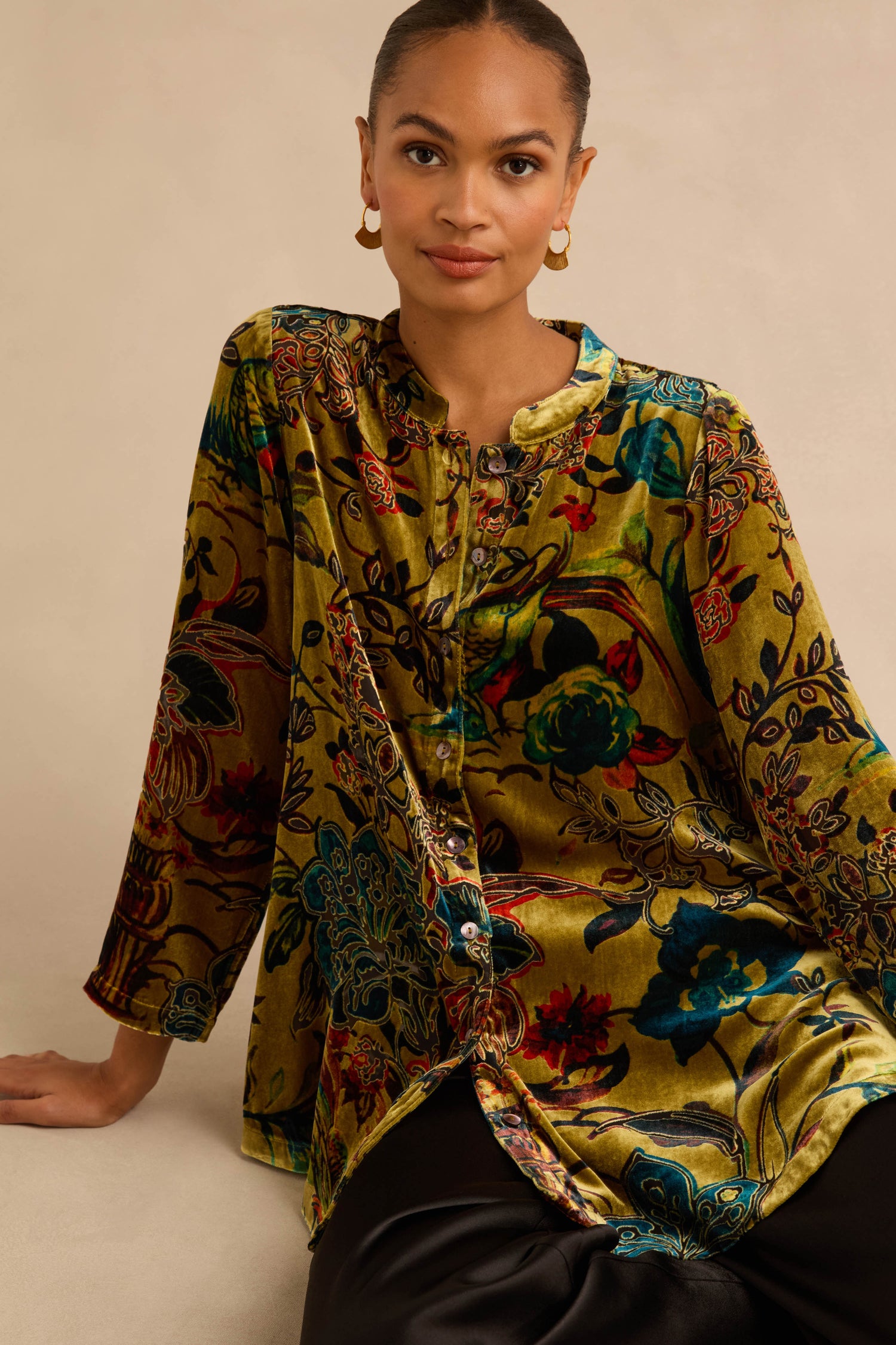 A person wearing a Silk Velvet Floral Devore Shirt, a long-sleeved, button-down top featuring a multi-colored floral pattern on olive-green fabric.